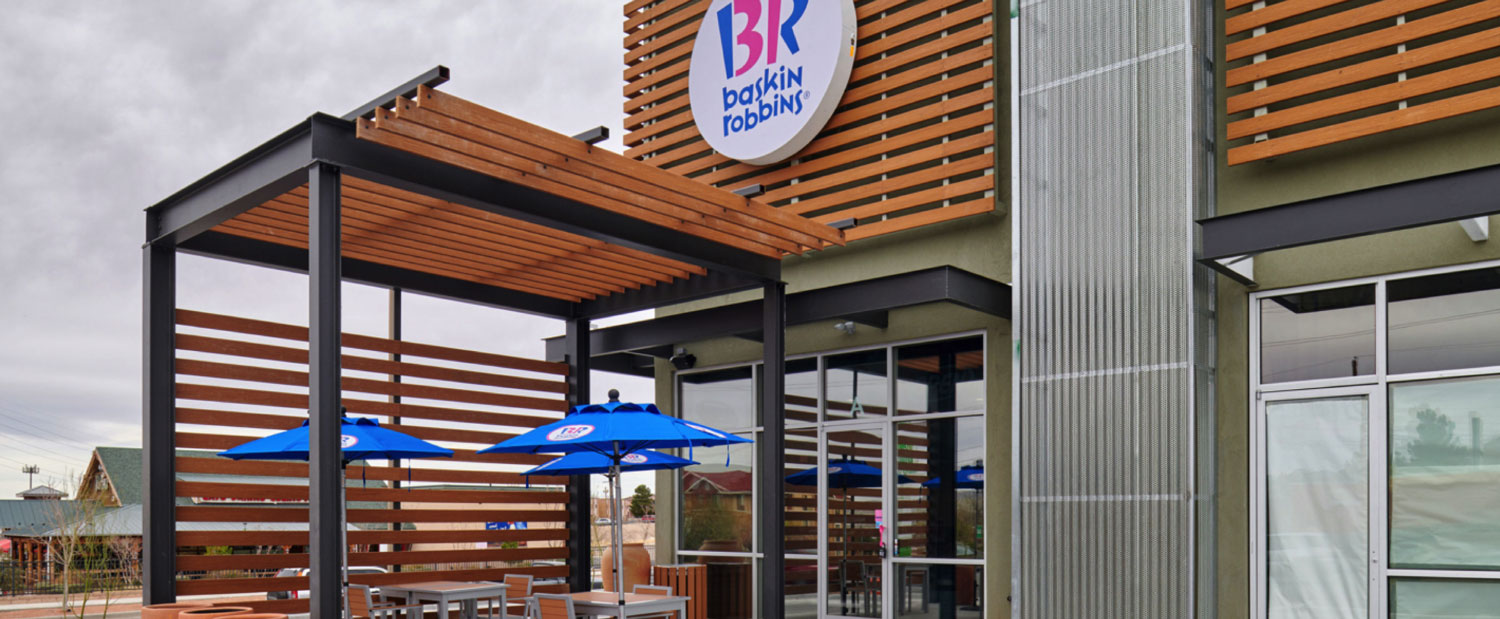 Exterior view of a baskin robbins store with a wooden façade and logo, featuring outdoor seating under blue umbrellas.