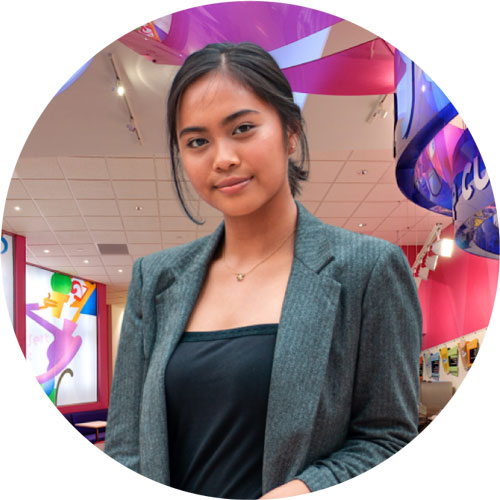 Young asian woman smiling in a grey blazer, standing inside a room with colorful decorations in the background.