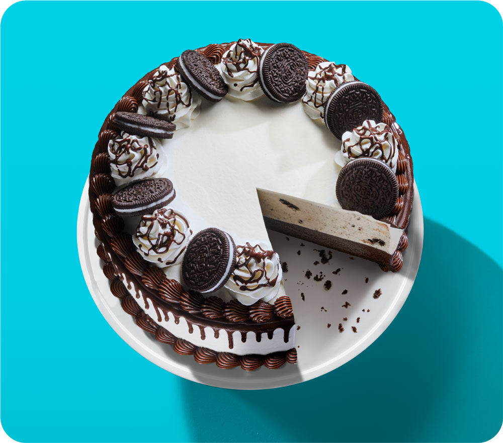 A round cake decorated with cream, chocolate drizzle, and whole and halved sandwich cookies, with one slice being lifted using a cake server.