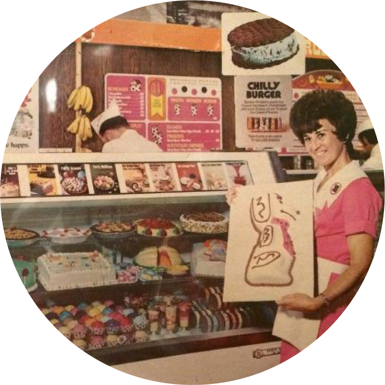 A woman in a pink uniform holds a menu beside an ice cream parlor counter filled with desserts and vintage advertisements on the wall.