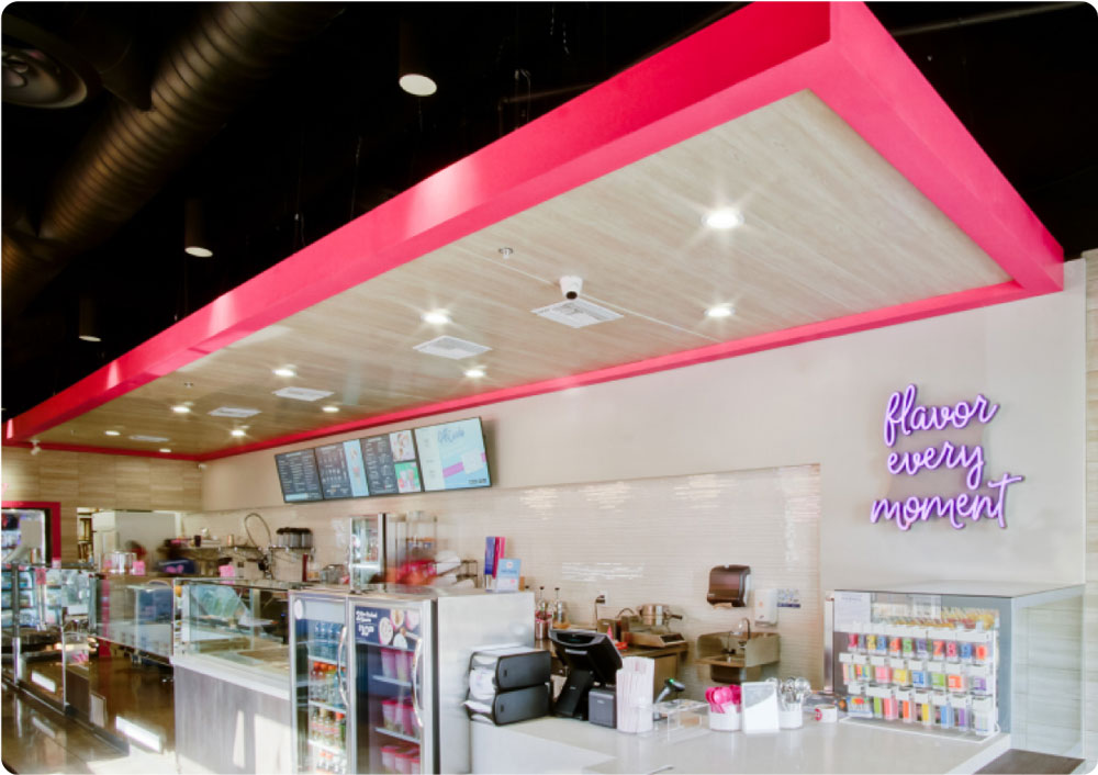 Interior of a modern bakery with a bright pink accent beam, wood paneling, glass display cases, and a neon sign that reads "flavor every moment.