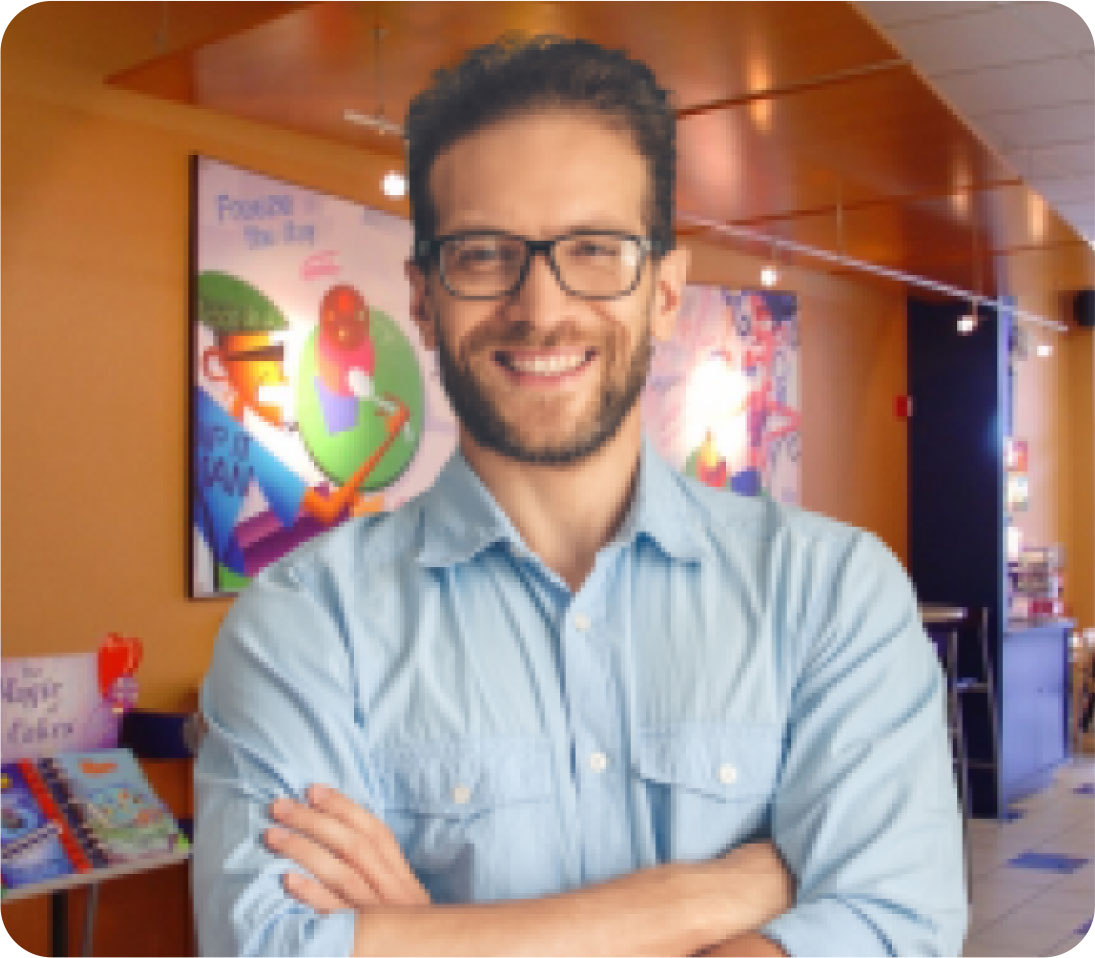 Smiling man with glasses and a blue shirt standing with arms crossed in a colorful bookstore.