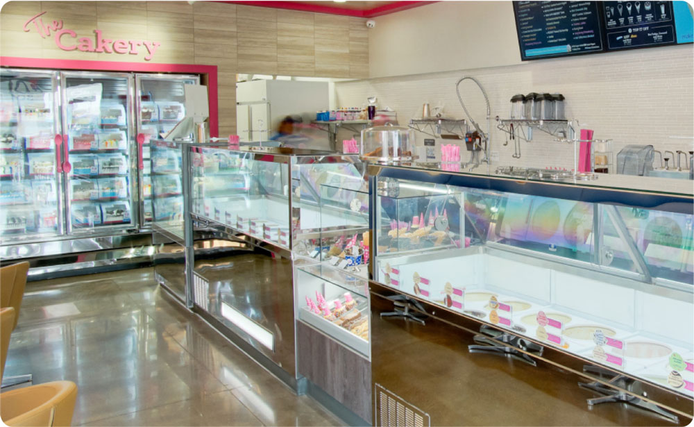 Interior of a modern bakery featuring glass display cases filled with assorted baked goods, refrigerators, and a menu board above the counter.